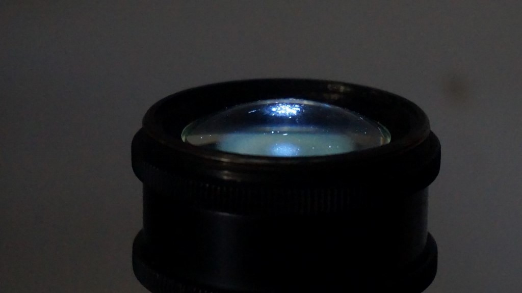 1.4 achromat collector lens being illuminated with an led flashlight from above and being viewed from an oblique angle. The illumination beam is just visible indicating that the maximum angle of acceptance is being reached. Image is deliberately upside down.