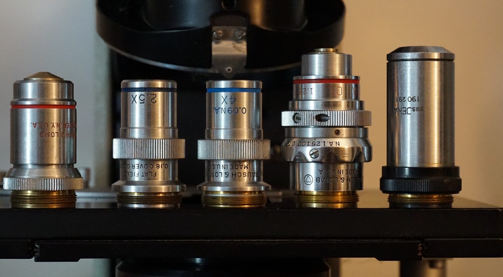 2.5X, 4X and 100X 1.25 iris planachromats, flanked by a conventional B &amp; L 97X 1.3 160mm objective on the left and a Jena D.I.N. objective on the right for comparison.